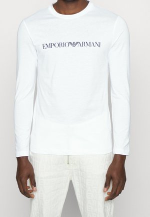 Mann trägt weißes Langarmshirt mit „EMPORIO ARMANI“-Logo in Marineblau, zentral auf der Brust.