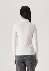 Long-sleeve ribbed top in light gray, features a collar and a slim fit. Fabric appears soft with a subtle texture. Worn with gray jeans.