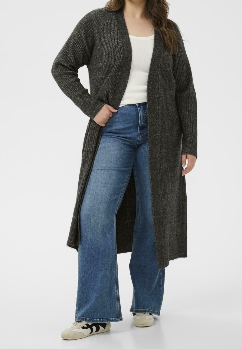 Femme portant un long cardigan côtelé gris foncé, un top blanc, un jean évasé bleu taille haute et des baskets blanches avec des bandes noires.