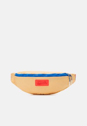 Beżowa nerka z niebieskim suwakiem i czerwonym naszywką z logo, wyposażona w podwójne suwaki i regulowane boczne paski.