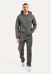 Felpa con zip verde oliva con tasche a marsupio e pantaloni abbinati. Presenta un piccolo logo e strisce laterali bianche sui pantaloni.