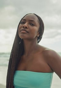 Portret van een persoon met lange gevlochten haren, gekleed in een turquoise strapless top, met een zachte, bewolkte strandachtergrond.