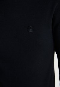 Sort ribbet strikket sweater med et subtilt broderet logo på brystet. Tæt siddende design med en glat tekstur.
