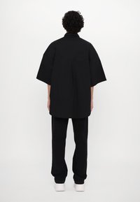 Black oversized shirt with wide sleeves and a pleat detail at the back, paired with black trousers and white shoes.