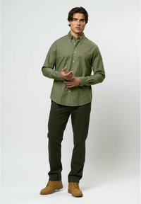 Cămașă cu butoni de culoare verde măslinie, cu o textură subtilă, asortată cu pantaloni verzi închisi și cizme bej. Cămașa are un mic logo roșu.