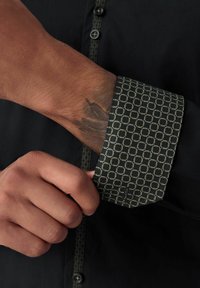 Maglietta nera con polsini a motivo che presenta disegni circolari in oro e blu. Il dettaglio dei bottoni è visibile nella zona del colletto.