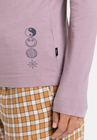 Camicia a maniche lunghe lavanda con disegni grafici che raffigurano icone celestiali e sorridenti. Indossata con pantaloni a quadretti gialli e bianchi.