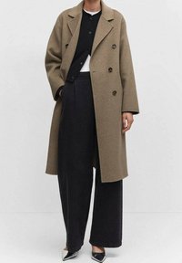 Cappotto di lana marrone doppiopetto con colletto, indossato sopra un cardigan nero e una maglietta bianca, abbinato a pantaloni neri a gamba larga e scarpe nere appuntite.