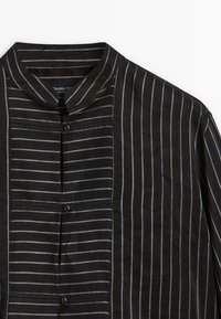 Camicia a strisce nere con colletto alla cinese. Caratterizzata da strisce orizzontali di larghezze diverse e chiusura con bottoni sul davanti. Tessuto leggero.