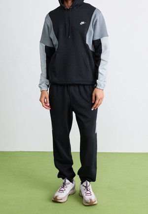 Person iført sort og grå Nike hoodie med matchende sorte Nike træningsbukser og hvide sneakers, stående på grøn gulv imod hvid væg.