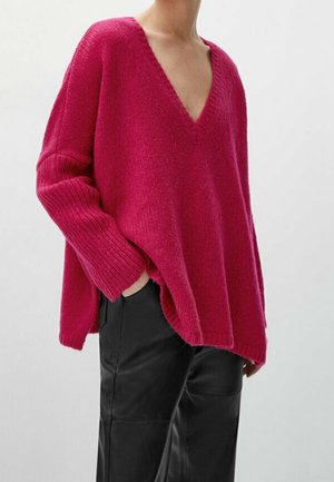 Woman wearing oversized deep pink V-neck sweater and black leather pants with one hand in pocket against plain background.