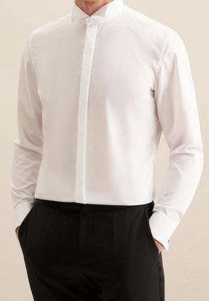 Mężczyzna noszący dopasowaną białą koszulę z ukrytymi guzikami i czarne eleganckie spodnie, ręce w kieszeniach, neutralne tło.