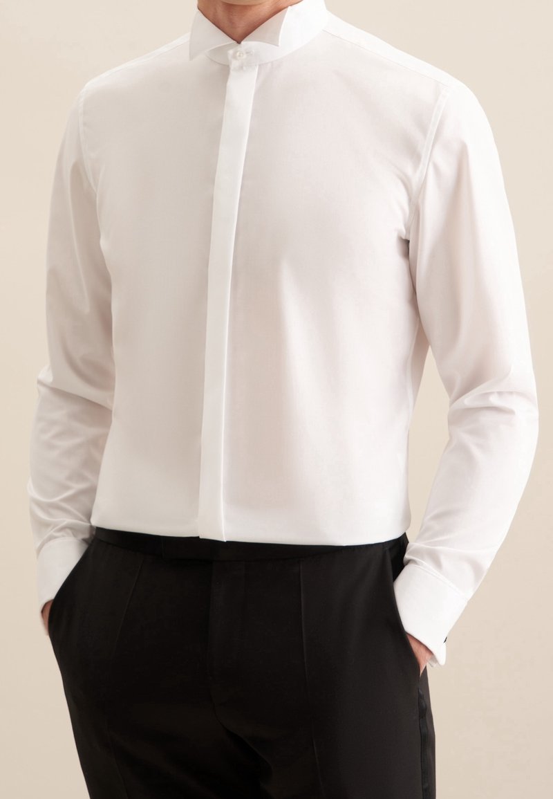 Homme portant une chemise blanche cintrée avec boutons cachés et un pantalon habillé noir, mains dans les poches, fond neutre.