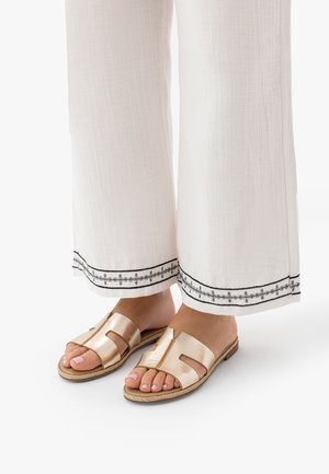 Picioare purtând sandale tip slider bej cu bretele late, stând alături, asortate cu pantaloni albi cu tivuri brodate negre.