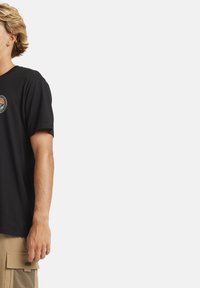 T-shirt in cotone nero con scollatura rotonda, maniche corte e una toppa grafica circolare sulla parte superiore sinistra del petto che presenta un design di montagna.