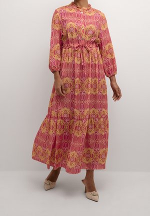 Kobieta w różowo-żółtej sukience z długimi rękawami sięgającej do kostek, nosząca beżowe szpilki z zaostrzonym noskiem.