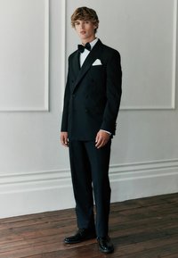 Schwarzer doppelt gesteppten Smoking mit satinierter Revers, weißem Hemd, schwarzer Fliege und Einstecktuch. Abgerundet mit polierten schwarzen Schuhen und taillierter Passform.