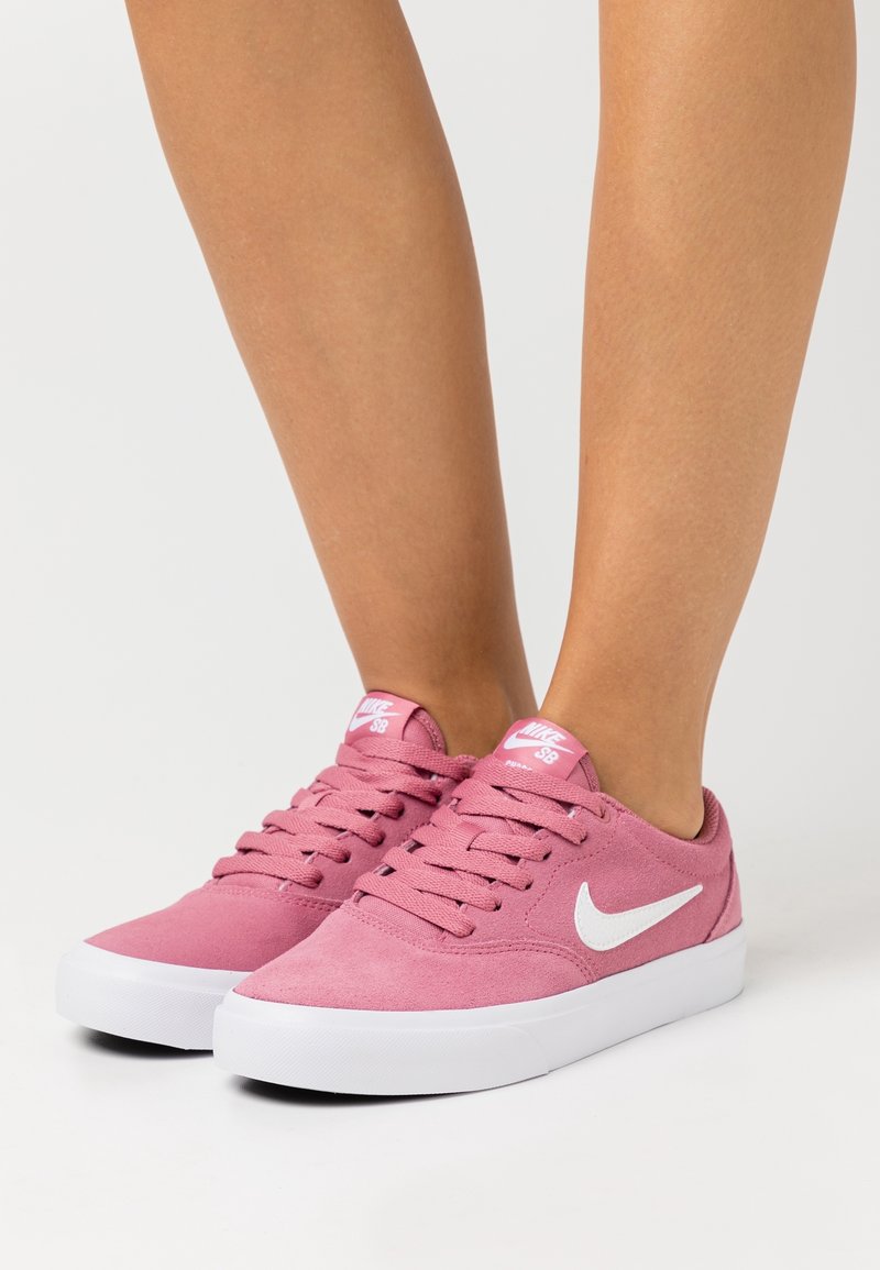 Jambes vêtues de baskets en daim rose Nike SB avec des logos Swoosh blancs et des semelles blanches, posées sur une surface gris clair.