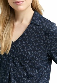 Marineblaue Bluse mit einem dezenten Wellenmuster in Hellblau und metallischen Akzenten, ausgestattet mit einem Reverskragen und einem weichen, strukturierten Stoff.