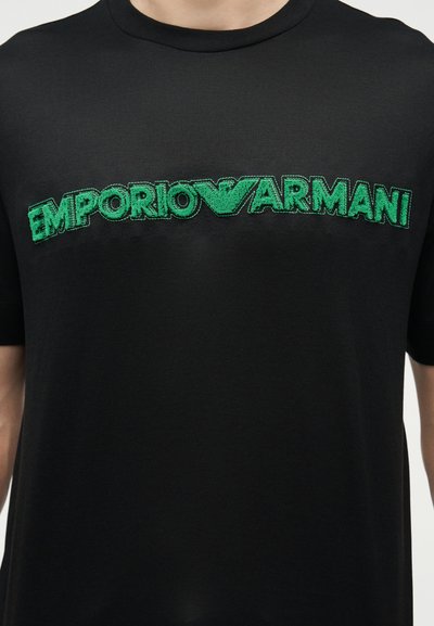 Μαύρο μπλουζάκι με πράσινο κεντημένο λογότυπο "EMPORIO ARMANI" στο στήθος, φορεμένο από ένα άτομο που φαίνεται από τον λαιμό μέχρι τη μέση.