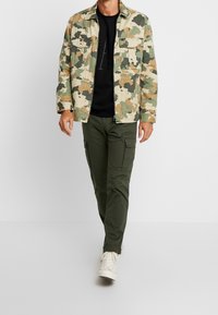 Chaqueta de camuflaje en tonos beige, verde y marrón, con dos bolsillos en el pecho, combinada con una camiseta negra y pantalones cargo verde olivo. Zapatillas blancas.