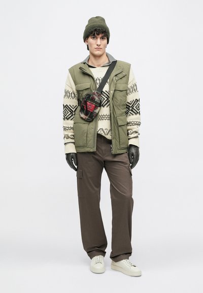 Λαδί φουσκωτό γιλέκο πάνω από ένα κρεμ πουλόβερ με σχέδια, καφέ cargo παντελόνι, μαύρα γάντια και μια καρό τσάντα χιαστί με κόκκινες λεπτομέρειες. Λευκά αθλητικά παπούτσια.