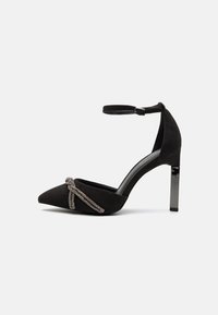 Chaussure à talon haut en suede noir avec un bout pointu, ornée d'un détail de nœud perlé et d'une bride de cheville. Talon noir brillant.