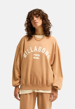 Vrouw met krullend haar die een beige Billabong-trui en bijpassende broek draagt, staand tegen een effen lichte achtergrond.