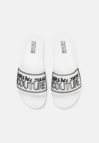 White slide sandals made of synthetic material, featuring a wide black band with "VERSACE JEANS COUTURE" printed in bold lettering.