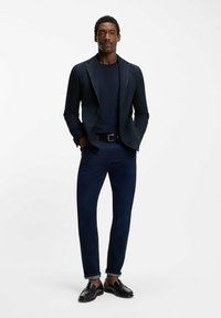 Mężczyzna w czarnej marynarce, granatowej koszuli, ciemnych dżinsach, czarnym pasku i czarnych mokasynach, stojący z rękami w kieszeniach na białym tle.