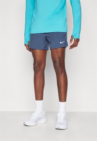 Nike shortsit tummansinisinä valkoisella logoilla, yhdistettynä turkoosiin pitkähihaiseen puseroon ja valkoisiin urheiluvaatteisiin, joissa on vaaleanharmaat yksityiskohdat.