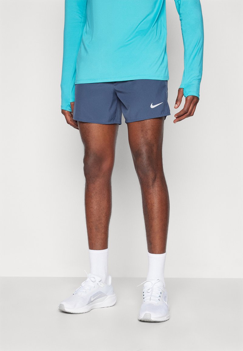 Nike shortsit tummansinisinä valkoisella logoilla, yhdistettynä turkoosiin pitkähihaiseen puseroon ja valkoisiin urheiluvaatteisiin, joissa on vaaleanharmaat yksityiskohdat.