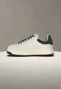 Λευκό sneaker χαμηλής κοπής με μαύρα κορδόνια, μαύρο επενδυμένο κολάρο φτέρνας και χοντρή λευκή σόλα με μαύρο πέλμα, εκτεθειμένο σε ουδέτερη επιφάνεια.