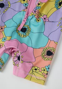 Maillot de bain floral coloré avec une fermeture éclair rose, présentant de grandes fleurs vives en violet, turquoise, jaune et orange sur fond pastel.