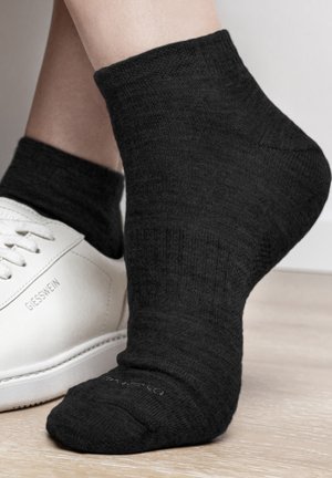 Schwarze Knöchelsocke aus Strickmaterial, mit einem gerippten Bund und glatter Textur. Getragen mit einem weißen Sneaker; Logo auf dem Schuh sichtbar.