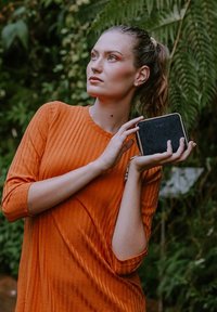 Black zippered wallet held in hand, featuring a smooth texture and gold hardware, paired with an orange ribbed long-sleeve top in natural settings.