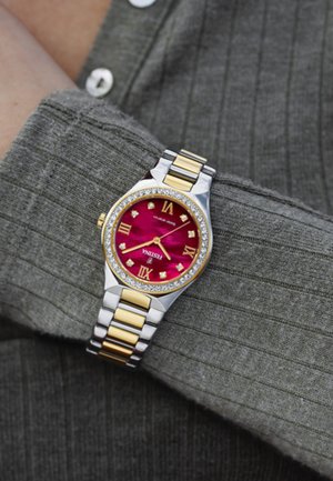 Reloj de oro y plata con una esfera roja vibrante, números romanos, adornos similares a diamantes y una pulsera de metal bicolor. Fondo multitético.