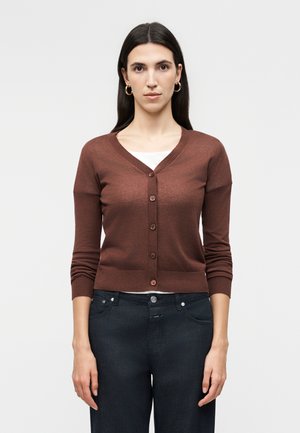 Kvinde med langt mørkt hår iført brun knaplukket cardigan, hvid top, mørke jeans og guld øreringe, stående foran en ensfarvet baggrund.