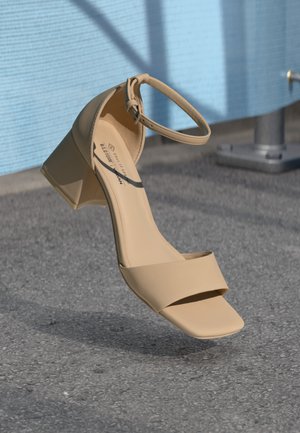 Sandale à talon carré beige avec un bout carré, des brides minimalistes et une boucle à la cheville. Matériau lisse et mat sur une surface grise.