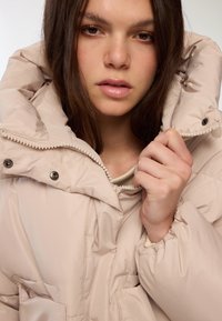 Kurtka puchowa w jasnym beżu, z wysokim kołnierzem, zamkiem błyskawicznym i zatrzaskami oraz widocznym przeszyciem. Miękka, gładka tkanina z pikowanym wzorem.