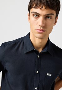 Νεαρός άνδρας με κοντά σκούρα μαλλιά που φοράει μαύρο πουκάμισο Wrangler με κουμπιά και γιακά, πάνω σε ένα απλό φωτεινό φόντο.