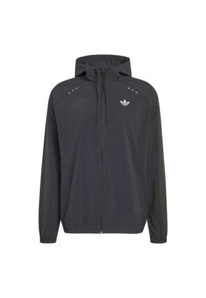 Jeune homme portant une veste à capuche noire zippée et un short noir, debout les mains dans les poches, regardant sur le côté.