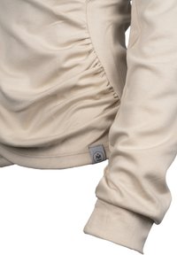 Beige pitkähihaine paita, jossa on ryppy yksityiskohta sivussa. Valmistettu pehmeästä puuvillakankaan ja varressa on hihansuuhun asti menevä sauma. Oikeassa alareunassa on vyöte.