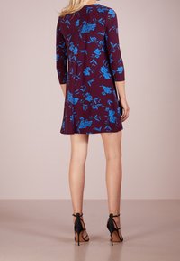 Lila klänning med blå blommönster, trekvartslånga ärmar, utsvängd design, knälängd, kombinerad med svarta remsandaler med hög klack.