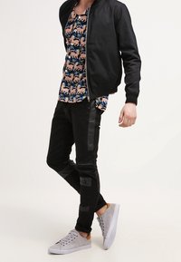 Chaqueta bomber negra sobre una camisa estampada con gatos rosas, combinada con unos jeans negros con paneles texturizados y zapatillas gris claro.