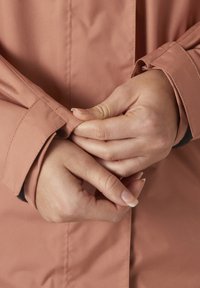 Chaqueta texturizada de color rosa claro con puños ajustados. Primer plano de manos ajustando las mangas, mostrando la suavidad del tejido y los detalles de la costura.