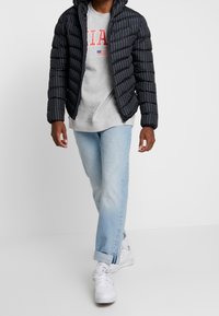Veste noire rembourrée avec des rayures horizontales, sweat-shirt gris en dessous, jeans bleu clair et baskets blanches. Design décontracté et moderne.