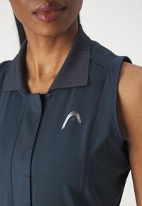 Femme portant une chemise sans manches de couleur bleu foncé avec un logo argenté sur la poitrine gauche, montrant le haut du torse et les lèvres.