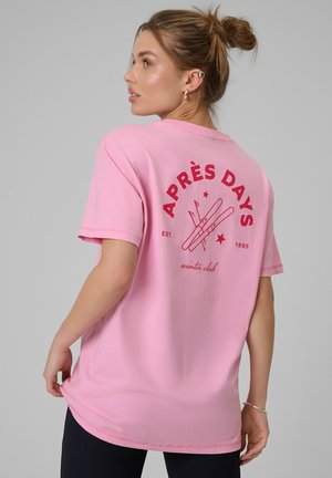 Młoda kobieta z włosami upiętymi w kok, ubrana w różowy t-shirt z czerwonymi nartami i napisem "APRÈS DAYS winter club" na plecach, na szarym tle.