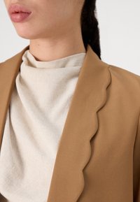 Gros plan sur une personne portant un blazer beige à bords festonnés par-dessus un haut crème clair texturé, avec le bas du visage et le cou visibles.